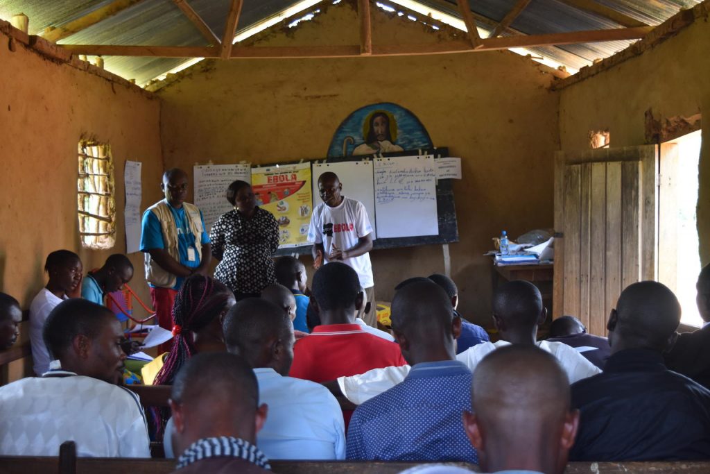 On 31 July 2018 in the Democratic Republic of the Congo, Leandre Kaseraka (right), an Ebola survivor, and UNICEF Communication Specialist Jean-Pierre Masuku (left) urge community members seated in a church in Mununze, North Kivu, to share toll-free numbers that provide information on the symptoms of Ebola.
Following the 1 August 2018 announcement by the Government of the Democratic Republic of the Congo (DRC) of a new Ebola Virus Disease (EVD) outbreak in North Kivu, UNICEF has mobilized its teams to help contain the spread of the disease and protect children. The impact of an outbreak on children can be far reaching. It’s known from earlier outbreaks in the DRC as well as in West Africa that children can be affected in various ways. Children can themselves be infected by the disease, but the impact goes beyond; it impacts their families and communities as children can lose their parents, care-givers and teachers. Access to basic services such as health care and education can become severely compromised. Also, children who are infected or whose relatives are, face stigmatization and social exclusion.
The Congolese Government has activated its response plan and called its partners, including UNICEF, to participate in the response. UNICEF has deployed a team to Beni for the response, including health specialists, communication specialists and a water, sanitation and hygiene specialist from the Ebola-response team in the Province of Equateur. Health, water, sanitation and hygiene and communication supplies have been sent to the affected areas including 300 laser thermometers to monitor the health conditions of people in the affected region and 2,000 kg of chlorine to treat water to help contain the spread of the disease. As at 14 August 2018, UNICEF has installed 35 chlorination points, as well as handwashing units in 45 public places and in health facilities in the affected areas of Beni and Mangina in North Kivu. UNICEF has supported the distribution of preven UNICEF/UN0231606/Herrmann